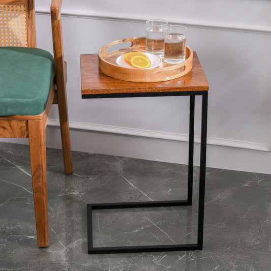 WOOD CURLS Odyssey C-shape side table with a warm mango wood top and black iron frame next to a green chair.