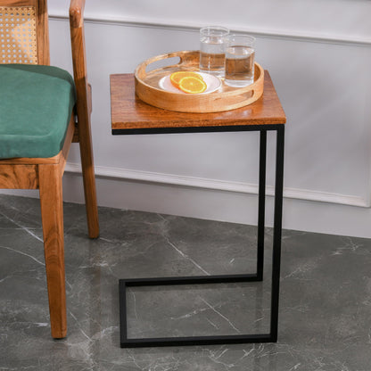 WOOD CURLS Odyssey C-shape side table with a warm mango wood top and black iron frame next to a green chair.
