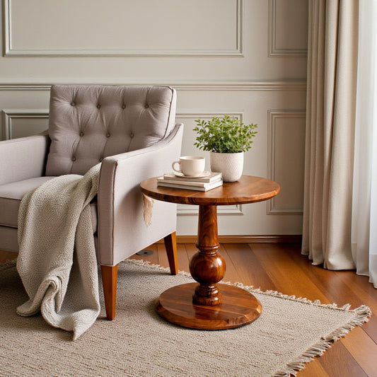 WOOD CURLS Elara round acacia wood side table with a pedestal base next to a beige upholstered armchair.