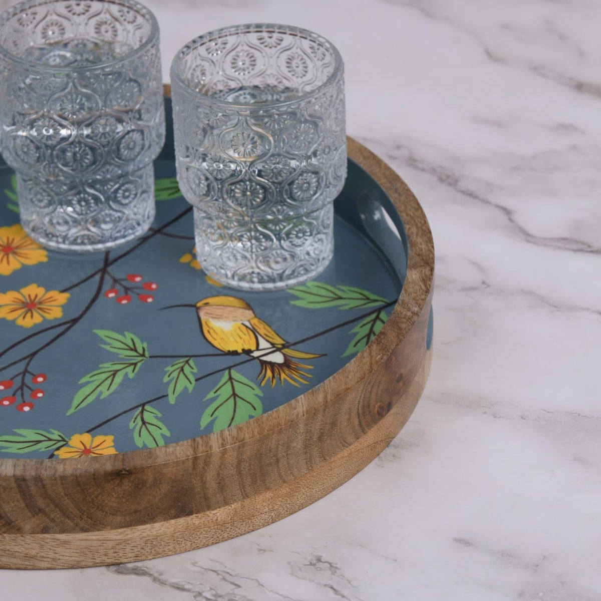 WOOD CURLS round mango wood tray with hand-painted bird and floral design on a slate blue background holding glasses
