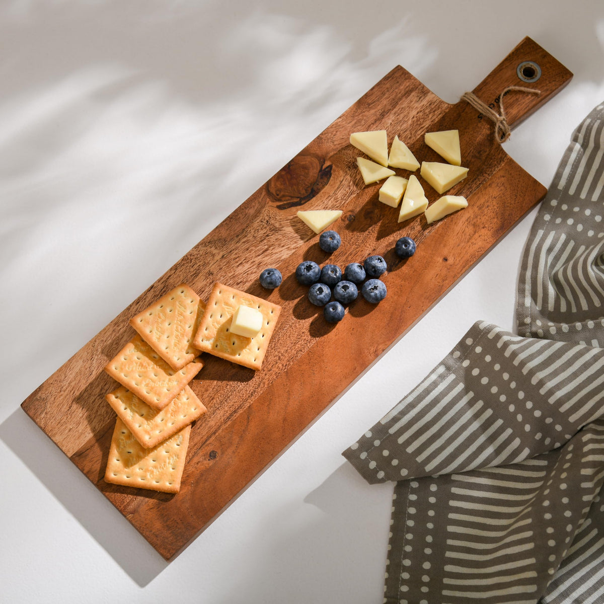 WOOD CURLS long acacia wood charcuterie board topped with crackers, cheese, and blueberries on a white surface.