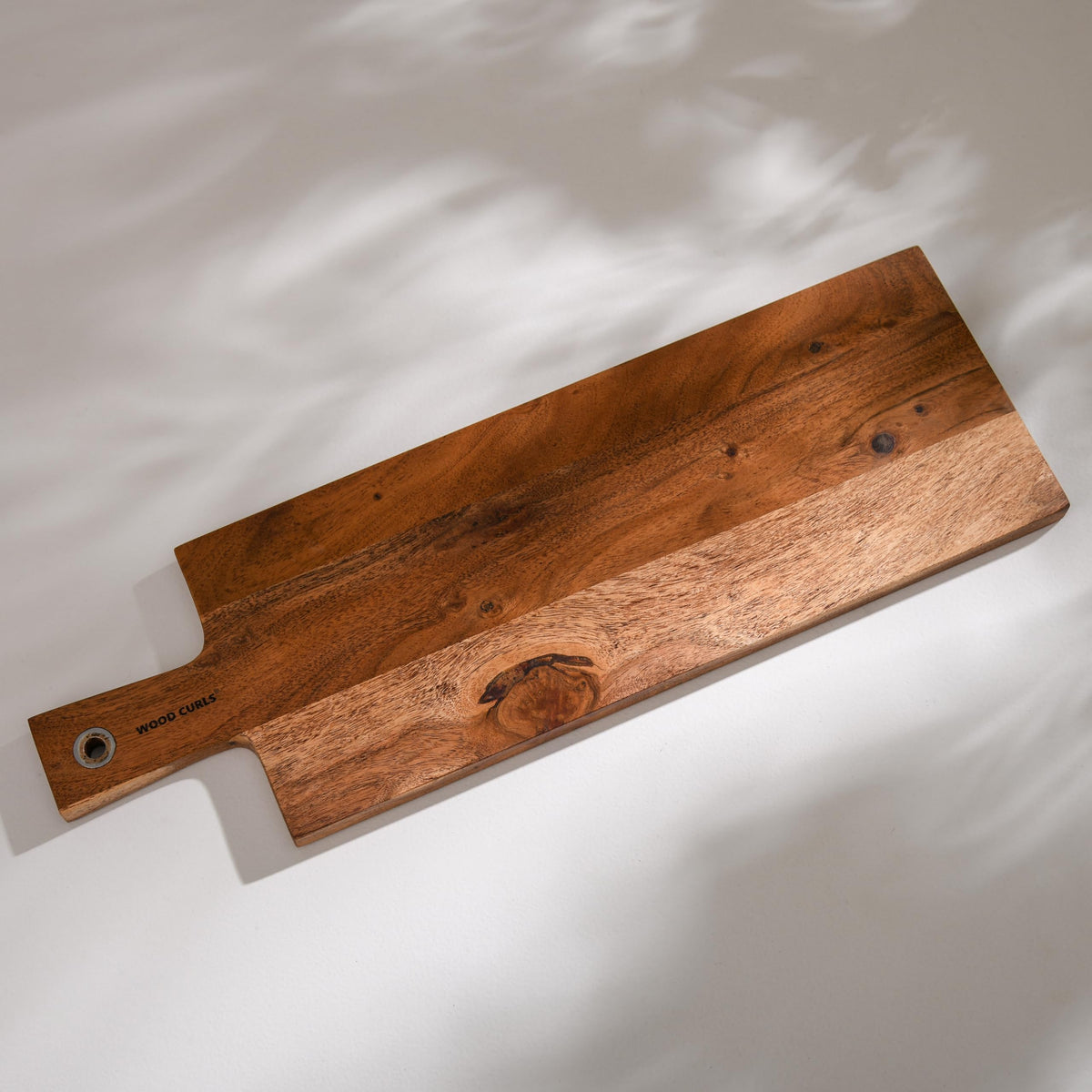 WOOD CURLS long acacia wood charcuterie board with a natural grain finish and integrated handle on a white surface.