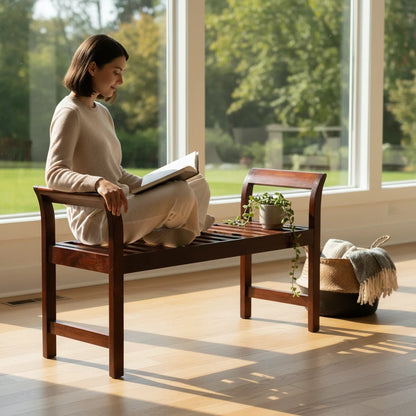 A woman reading on a WOOD CURLS solid wood bench with a mahogany finish, slatted seat, and arms in a sunlit room.