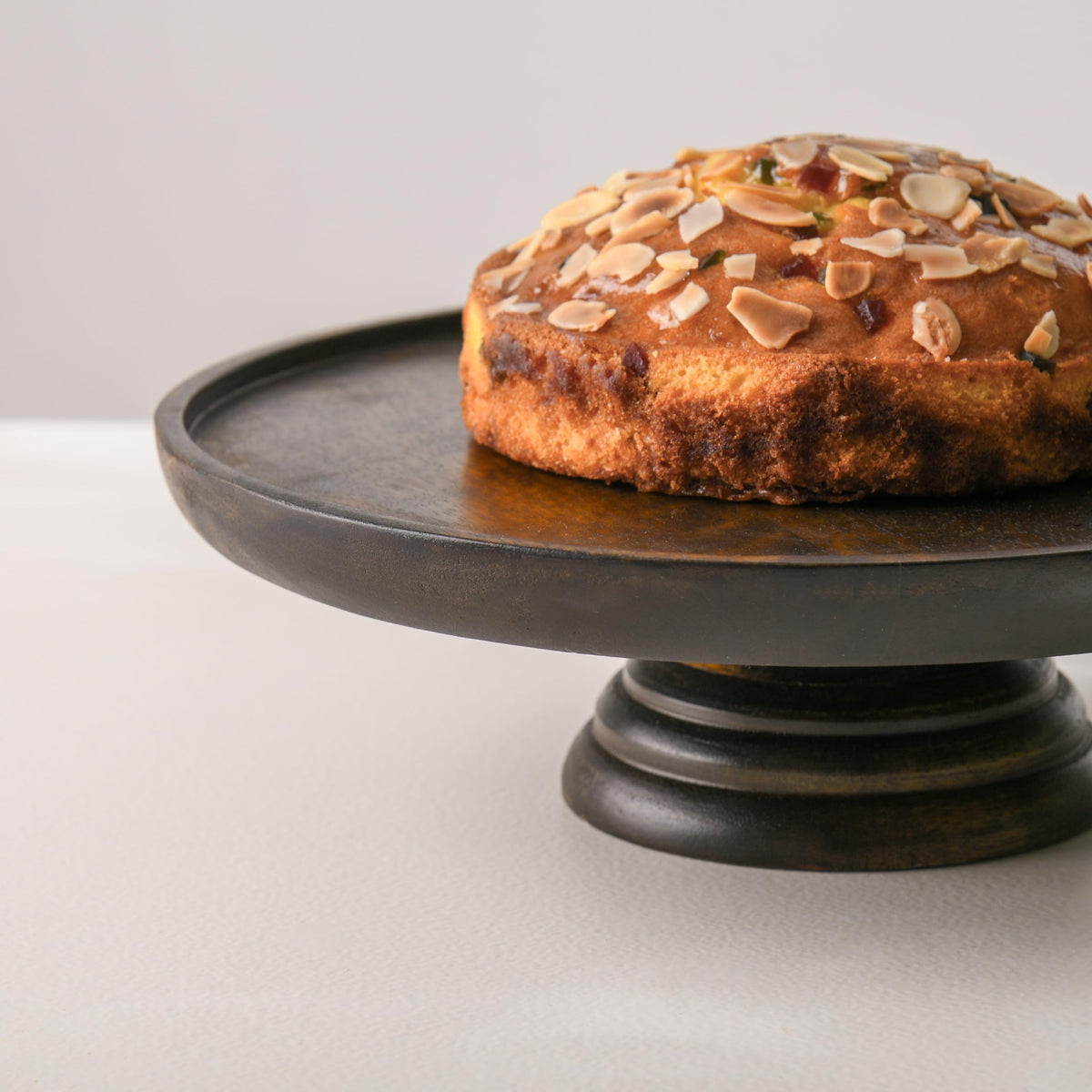 WOOD CURLS round mango wood pedestal cake stand with a walnut finish displaying a cake topped with almond slices.