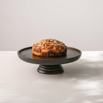 WOOD CURLS round mango wood pedestal cake stand with a rich walnut finish displaying a cake topped with almonds.