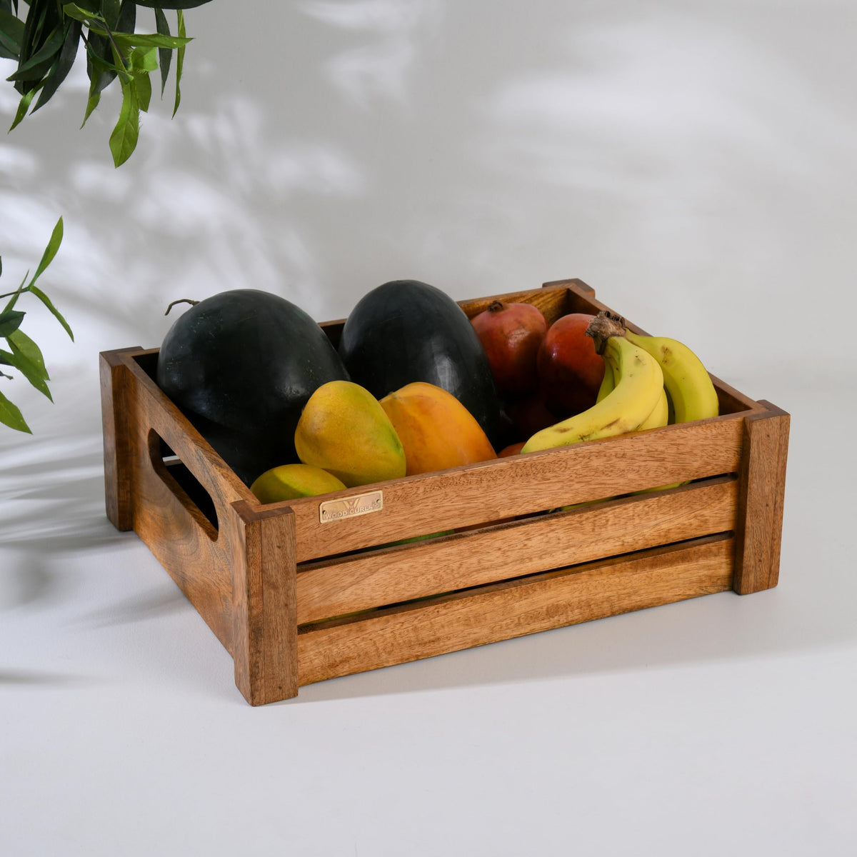 WOOD CURLS handcrafted mango wood crate filled with fresh fruits like bananas, pomegranates, and papayas on a white table.