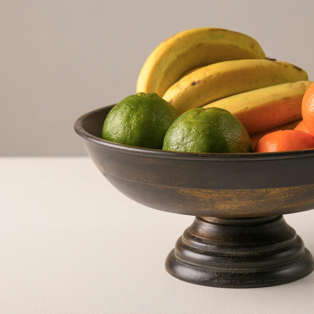 WOOD CURLS mango wood pedestal bowl with a walnut finish, filled with fresh fruit on a neutral table surface.