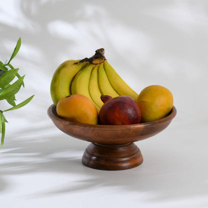 WOOD CURLS handcrafted mango wood pedestal fruit bowl filled with fresh bananas and mangoes on a white surface.