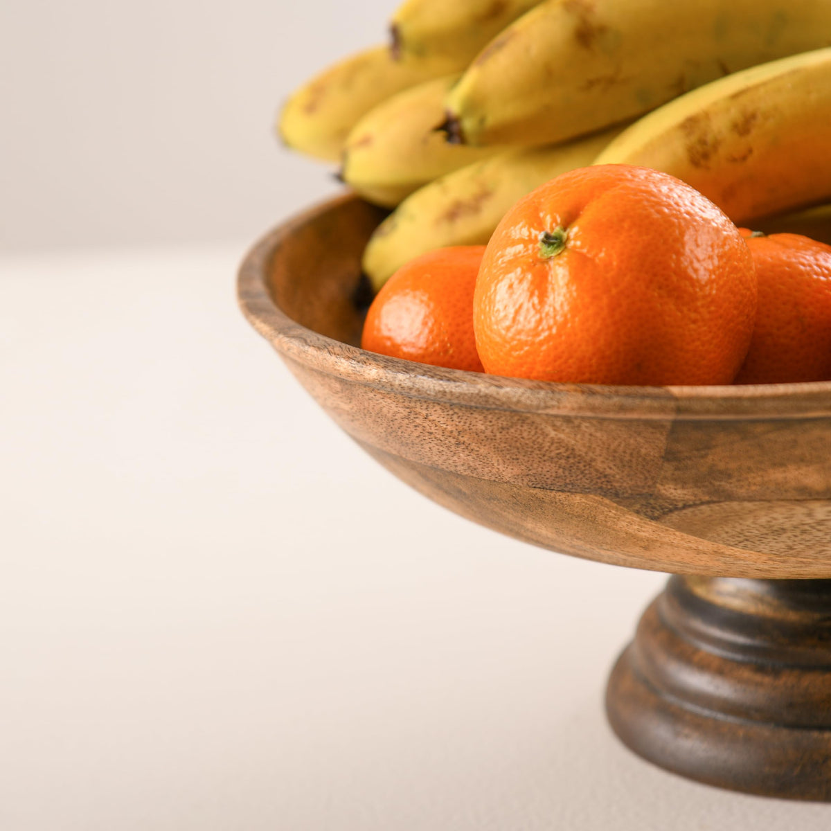 WOOD CURLS handcrafted mango wood pedestal bowl filled with fresh bananas and oranges on a neutral surface