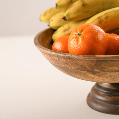 WOOD CURLS handcrafted mango wood pedestal bowl filled with fresh bananas and oranges on a neutral surface