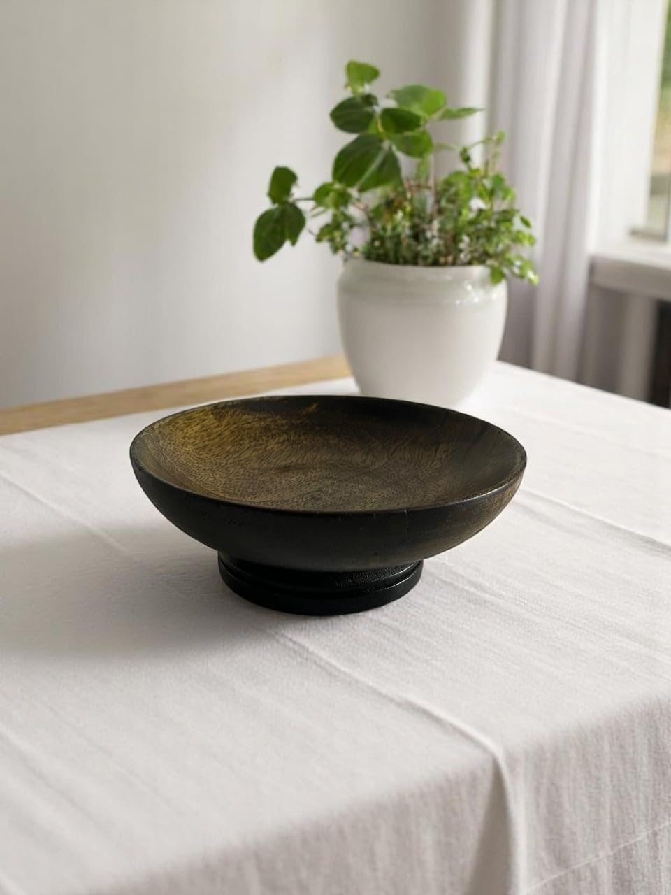 WOOD CURLS small dark brown mango wood pedestal bowl on a white tablecloth with a potted plant in the background