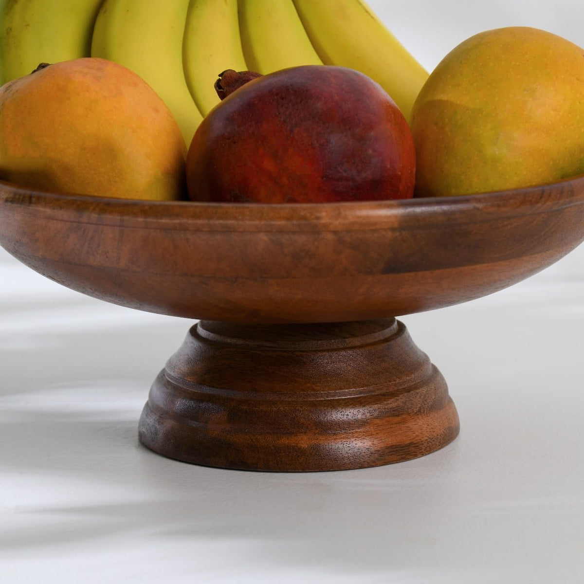 WOOD CURLS handcrafted mango wood pedestal fruit bowl filled with fresh bananas, mangoes, and a pomegranate.