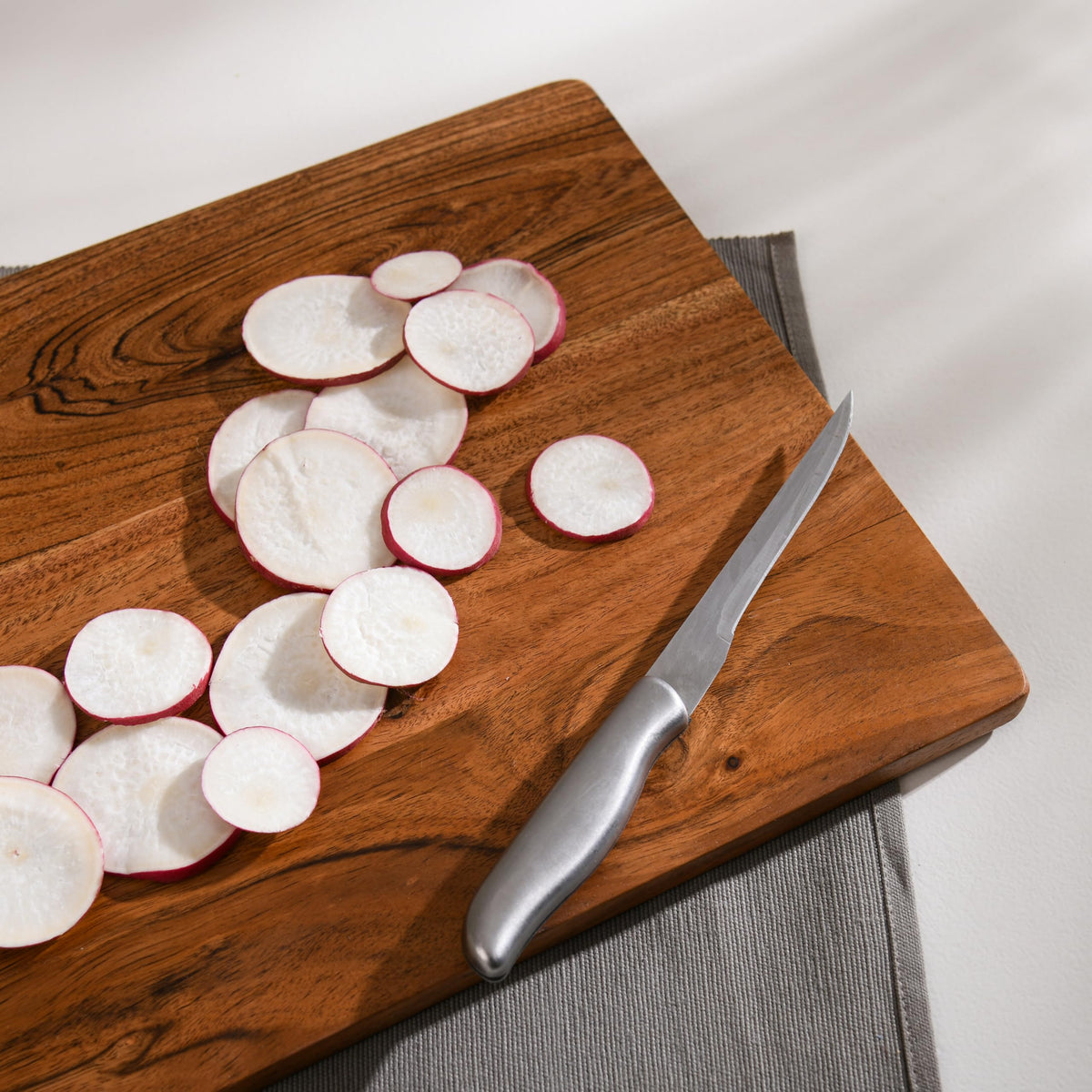 WOOD CURLS rectangular wooden chopping board with sliced radishes and a stainless steel knife on a grey placemat