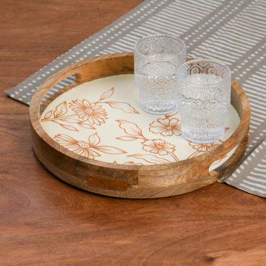 WOOD CURLS round mango wood serving tray with cream base and copper floral design, holding two textured glass tumblers.