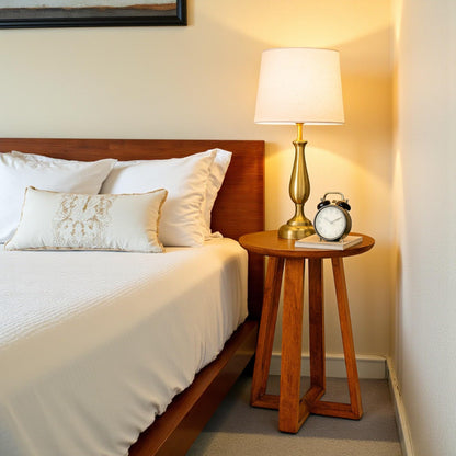 WOOD CURLS Mirage round mango wood side table with natural finish placed next to a bed with a lamp and alarm clock.