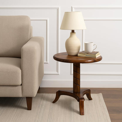 WOOD CURLS round end table in natural Sheesham wood with a pedestal base, styled with a lamp and mug next to a sofa.