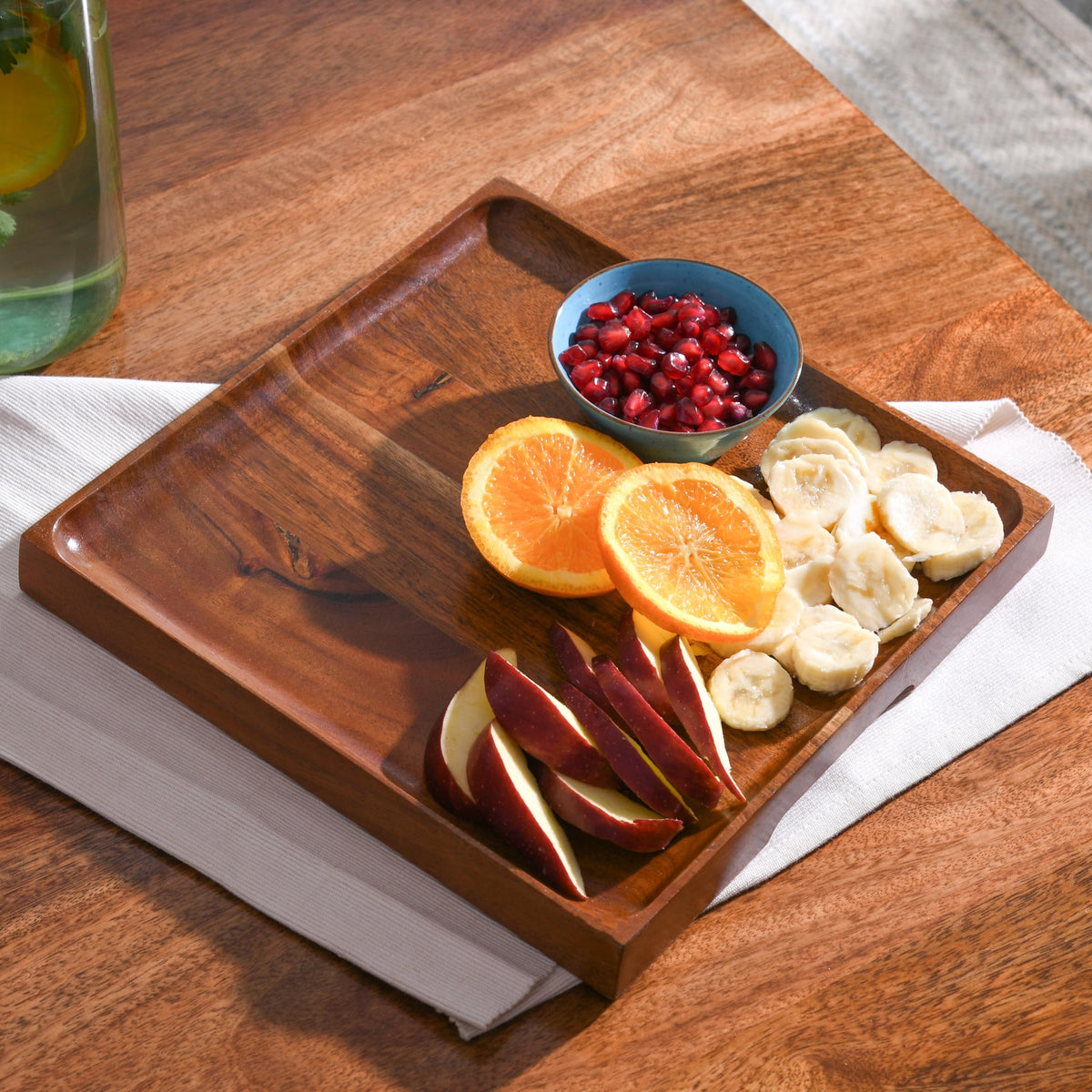 WOOD CURLS square mango wood serving platter holding sliced oranges, bananas, apple wedges, and pomegranate seeds.